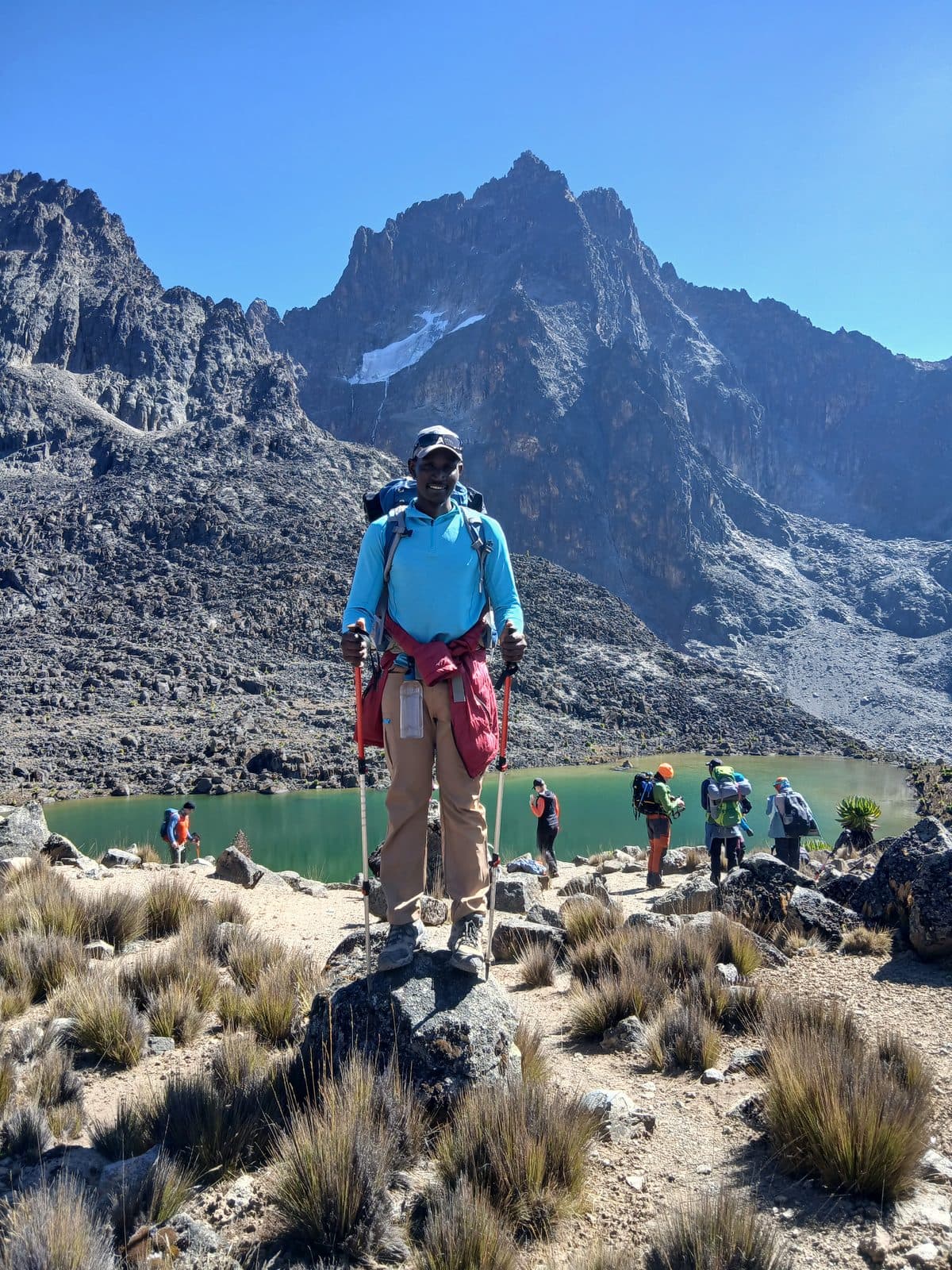 Guide in a blue shirt standing on rocks above a green alpine lake below Mount Kenya