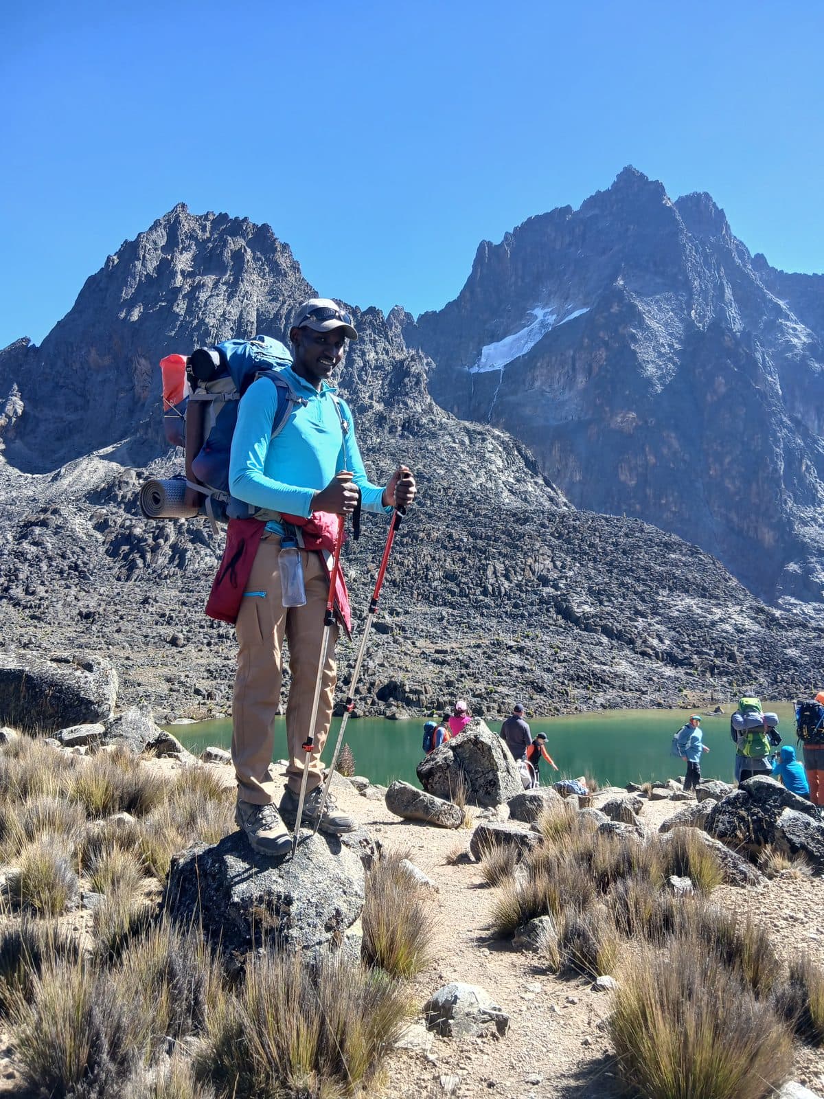 Guide with trekking poles beside a green alpine tarn under Mount Kenya's peaks